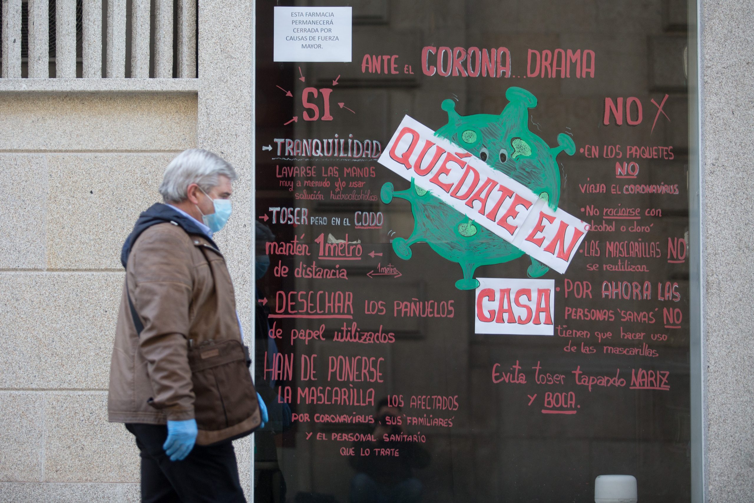 un hombre con mascarilla y guantes pasa al lado de un escaparate de una tienda con el mensaje de "quédate en casa"